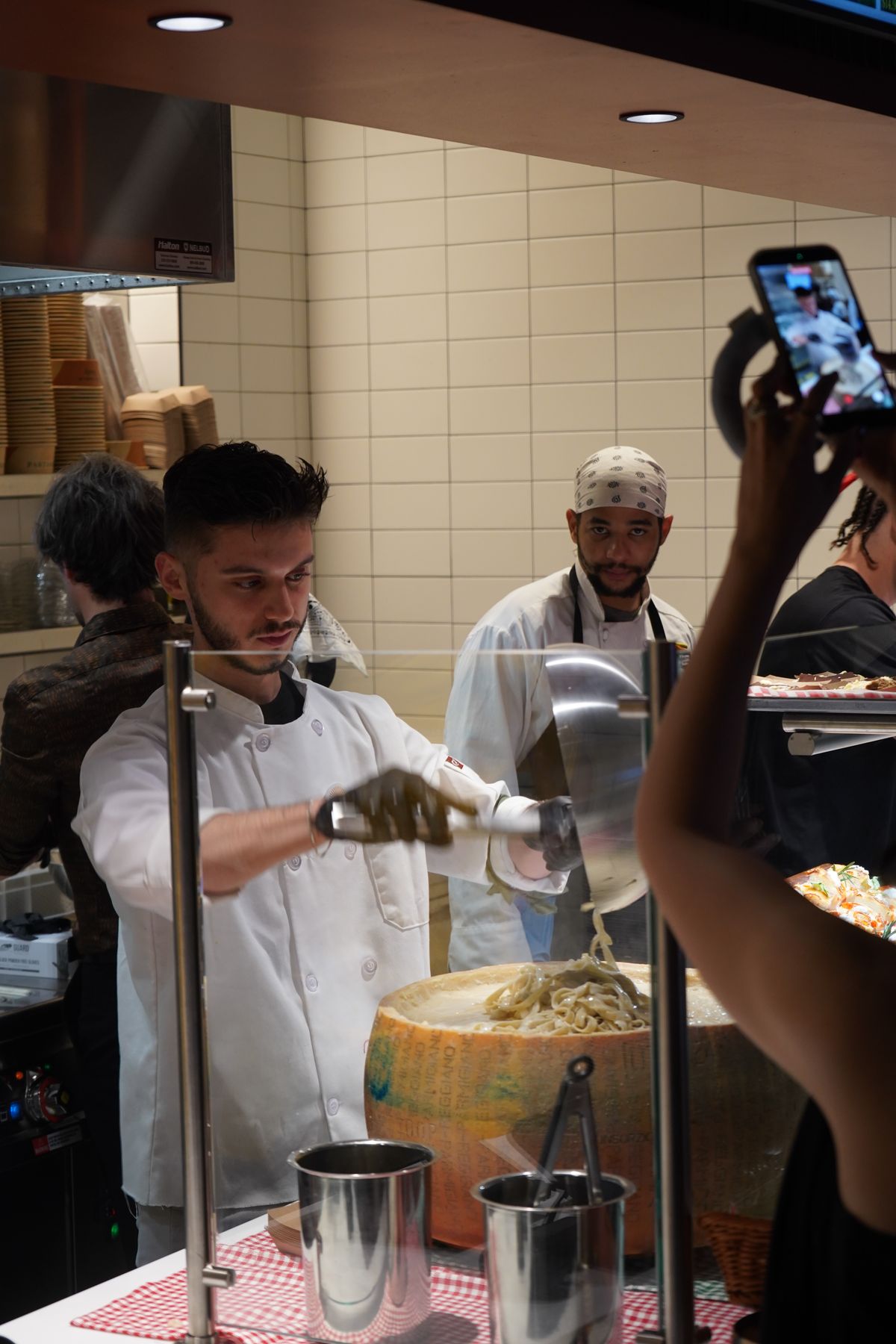Pastasole chef tossing fresh fettuccine in a Parmigiano Reggiano wheel while a guest films it on a phone